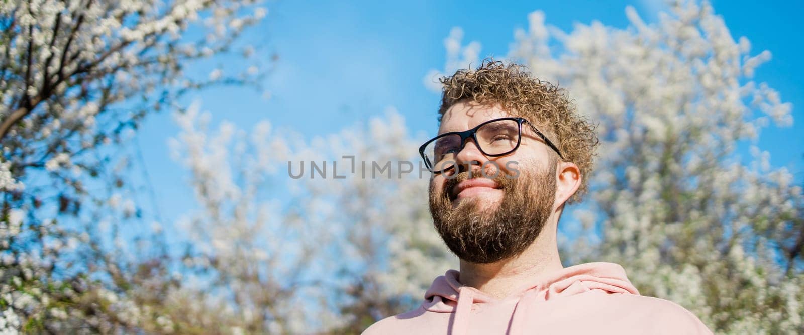 Male bearded guy standing under branches with flowers of blooming almond or cherry tree in spring garden. Spring blossom. Copy space