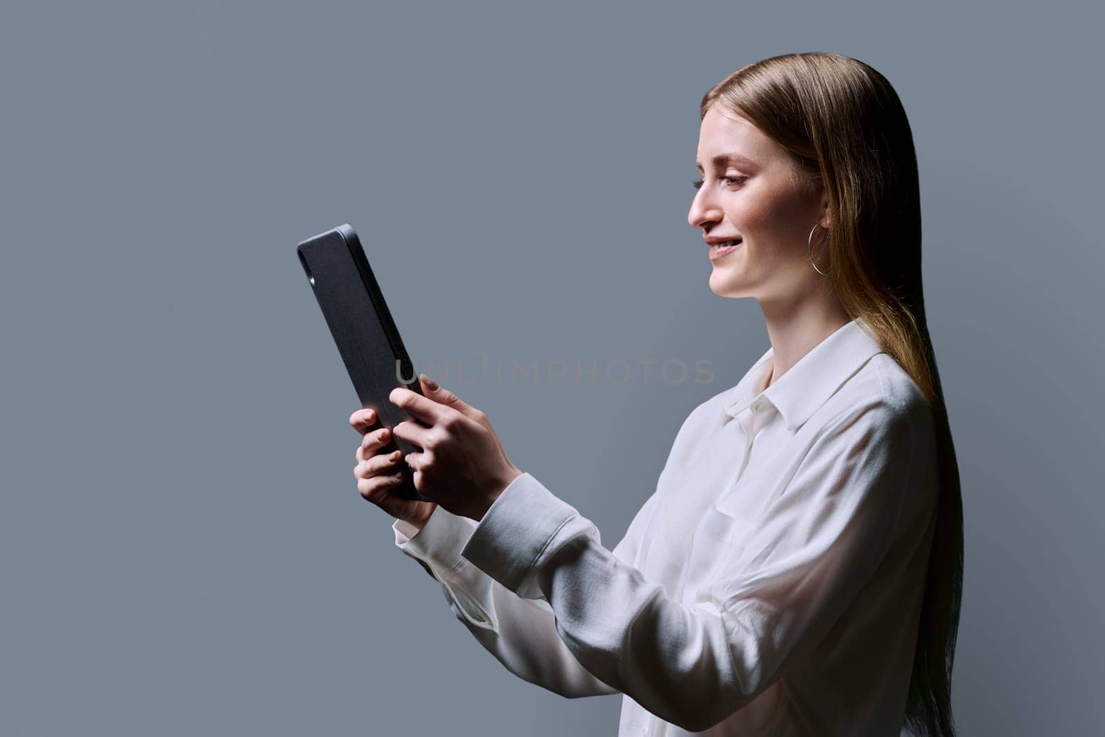 Teenage female student using digital tablet on gray background, profile view. Teenager girl 16,17, 18 years old reading looking at screen. Education Internet technology e-learning educational services