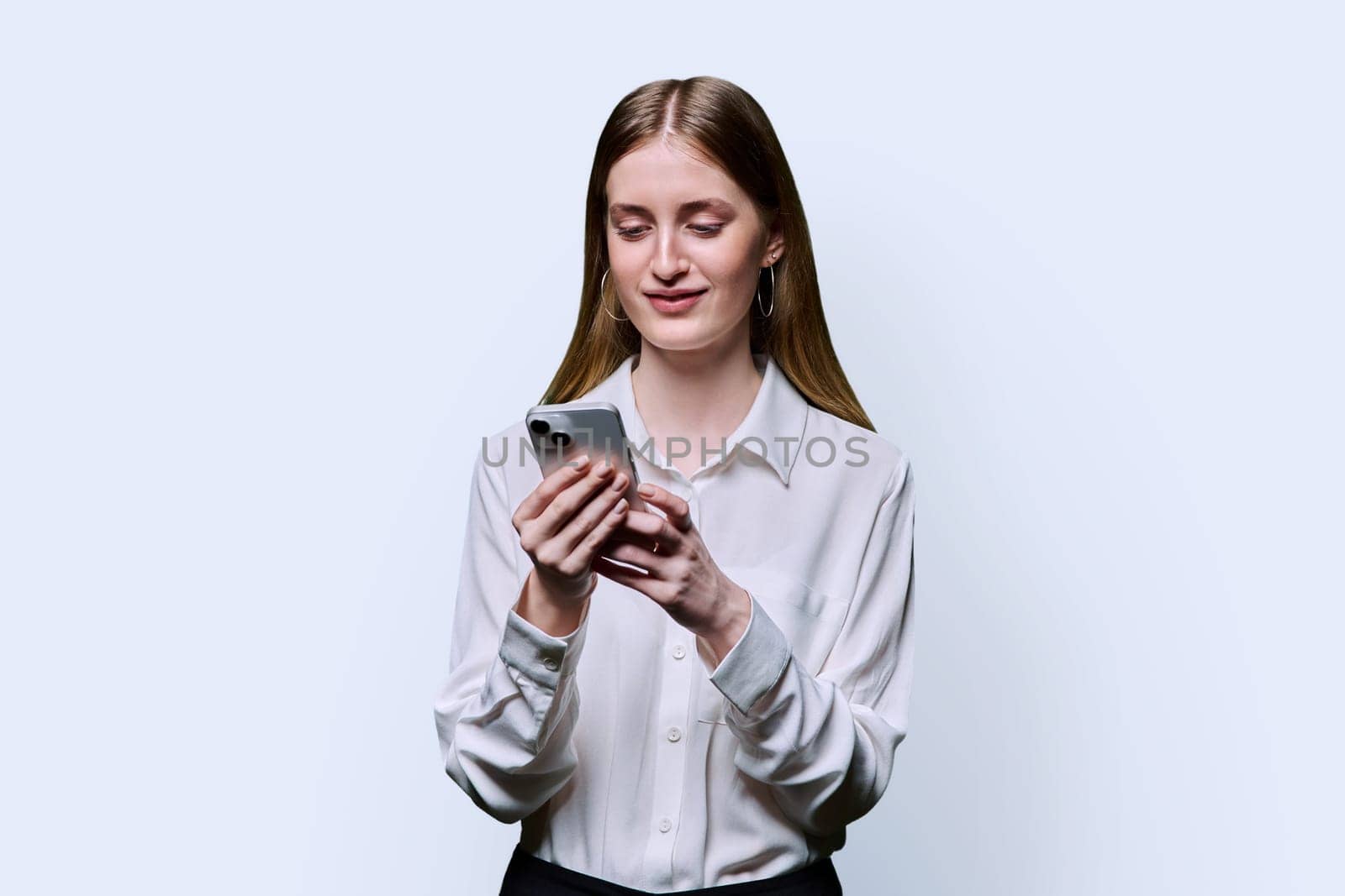 Teenage student girl 16, 17, 18 years old in white shirt holding smartphone in hands, texting looking at screen on white studio background. Education technology high school college lifestyle youth