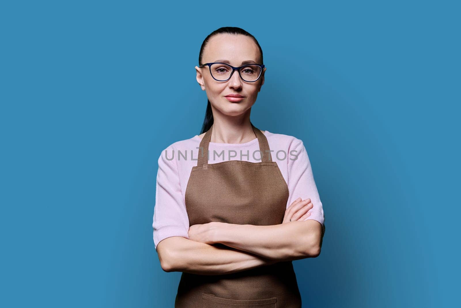 Portrait of confident 30s woman in apron on blue studio background. Successful female small business owner, service worker looking at camera with crossed arms. Staff, management, advertising, people