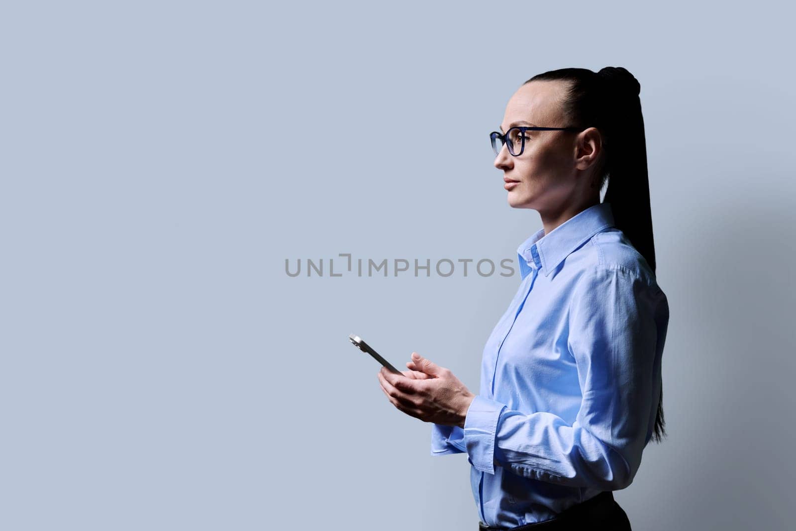 Profile view 30s serious woman using smartphone on grey background. Middle aged female in glasses looking forward. Technologies mobile apps applications internet work business leisure communication