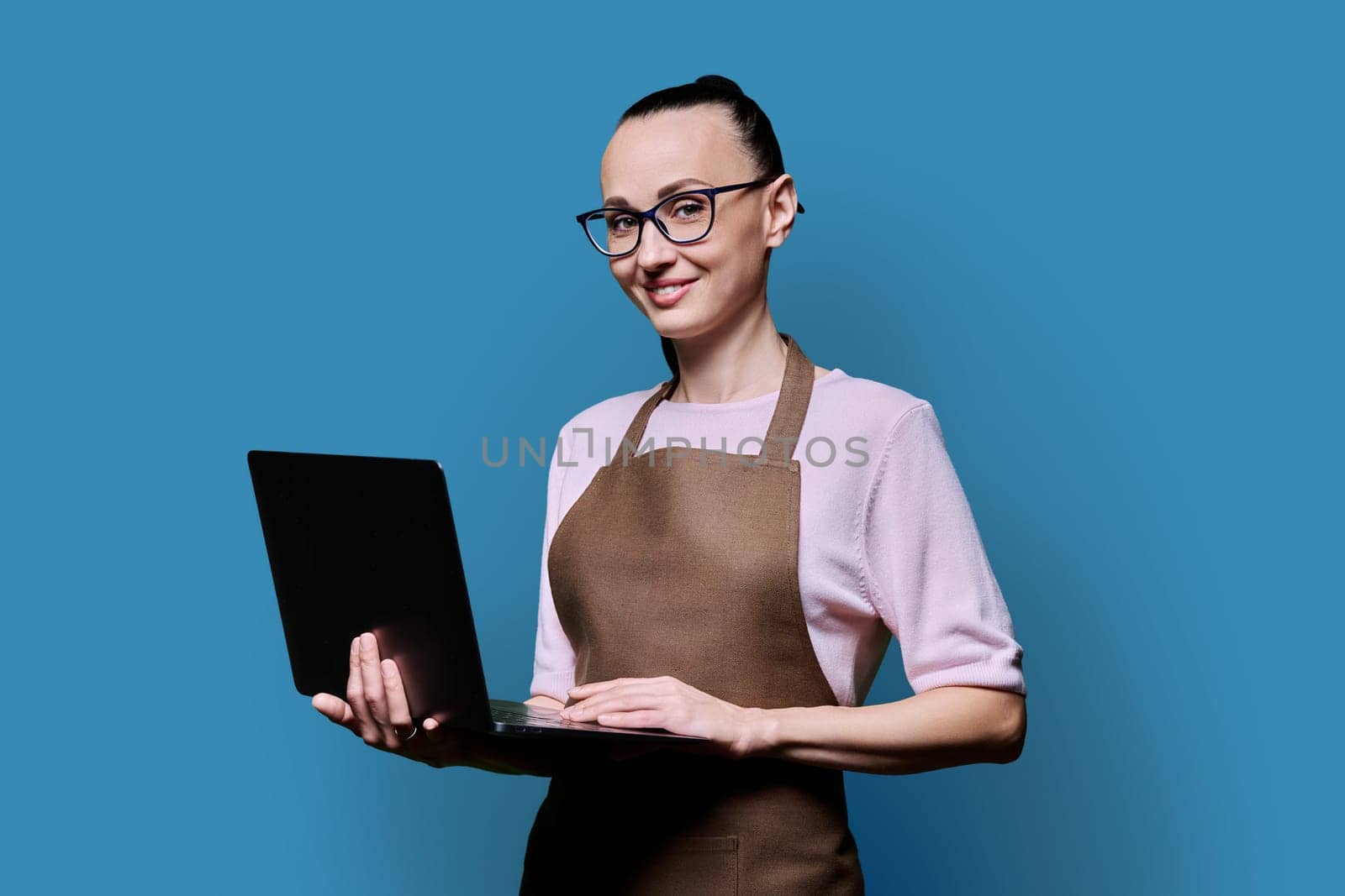 Portrait of 30s smiling confident woman in an apron using laptop, looking at camera on blue studio background. Worker, startup, small business, job, service sector, staff, youth concept