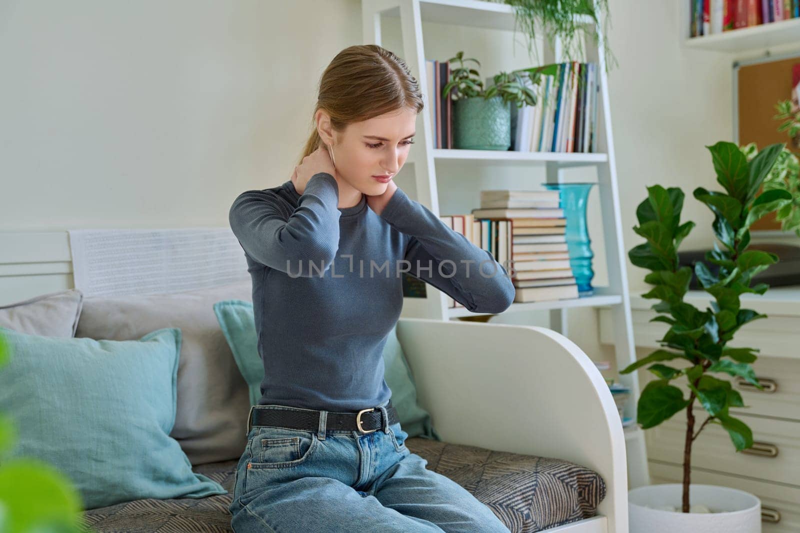 Young teenage female experiencing neck pain sitting on couch at home by VH-studio