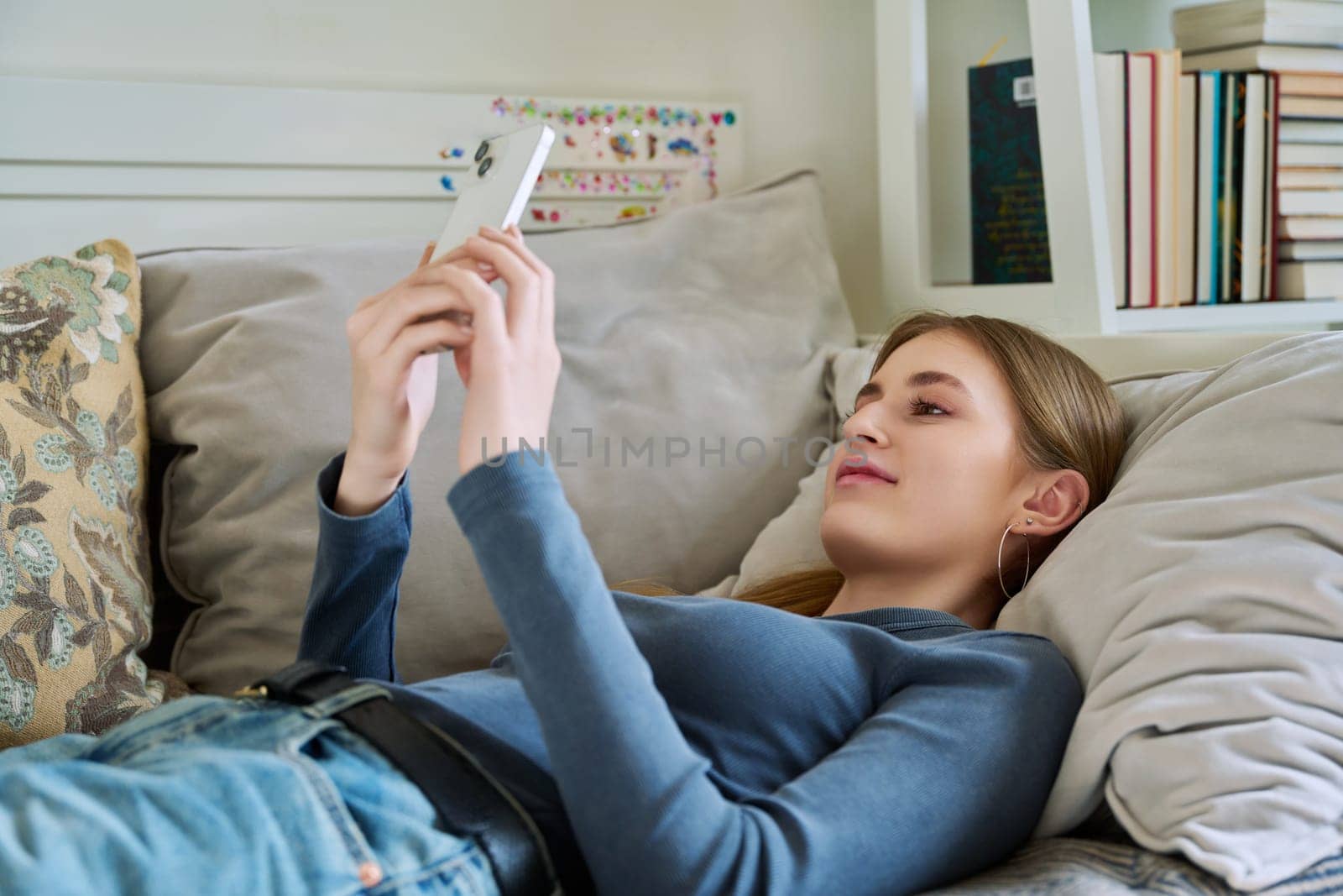 Happy relaxed female teenager lying on couch at home using smartphone by VH-studio