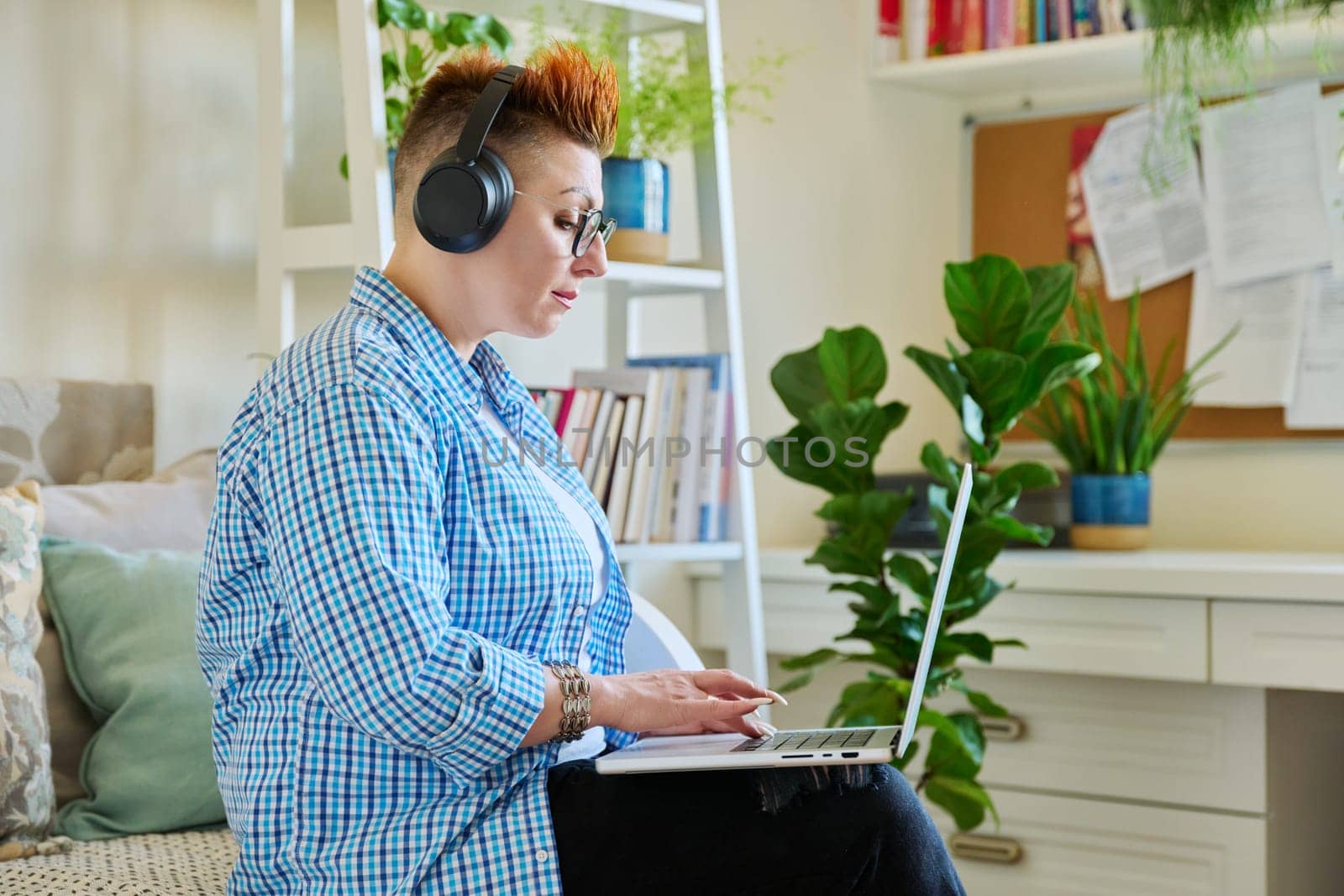 Middle aged woman freelancer in headphones using laptop computer sitting on couch at home. 40s female looking at screen video chat conference call online meeting training mentoring technology for work