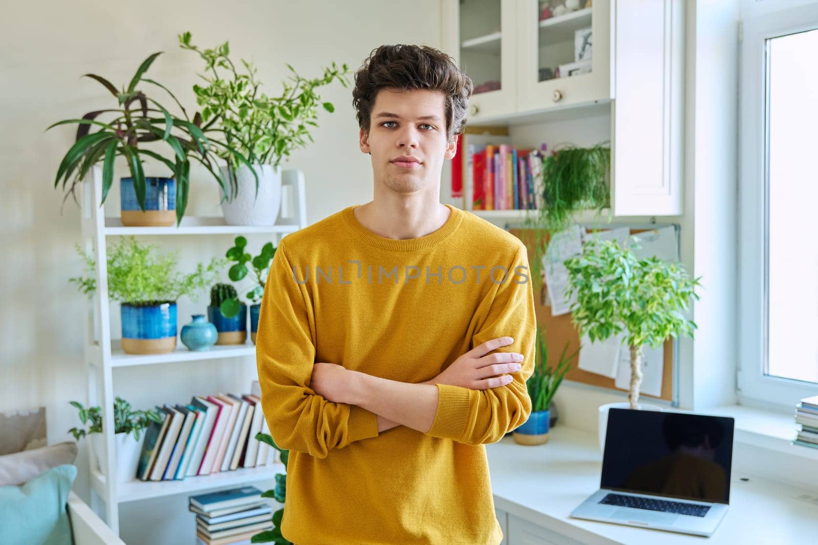 Portrait of young handsome guy with crossed arms, in home interior. Smiling confident male 19-20 years old in casual yellow with curly hairstyle looking at camera. Lifestyle, youth concept