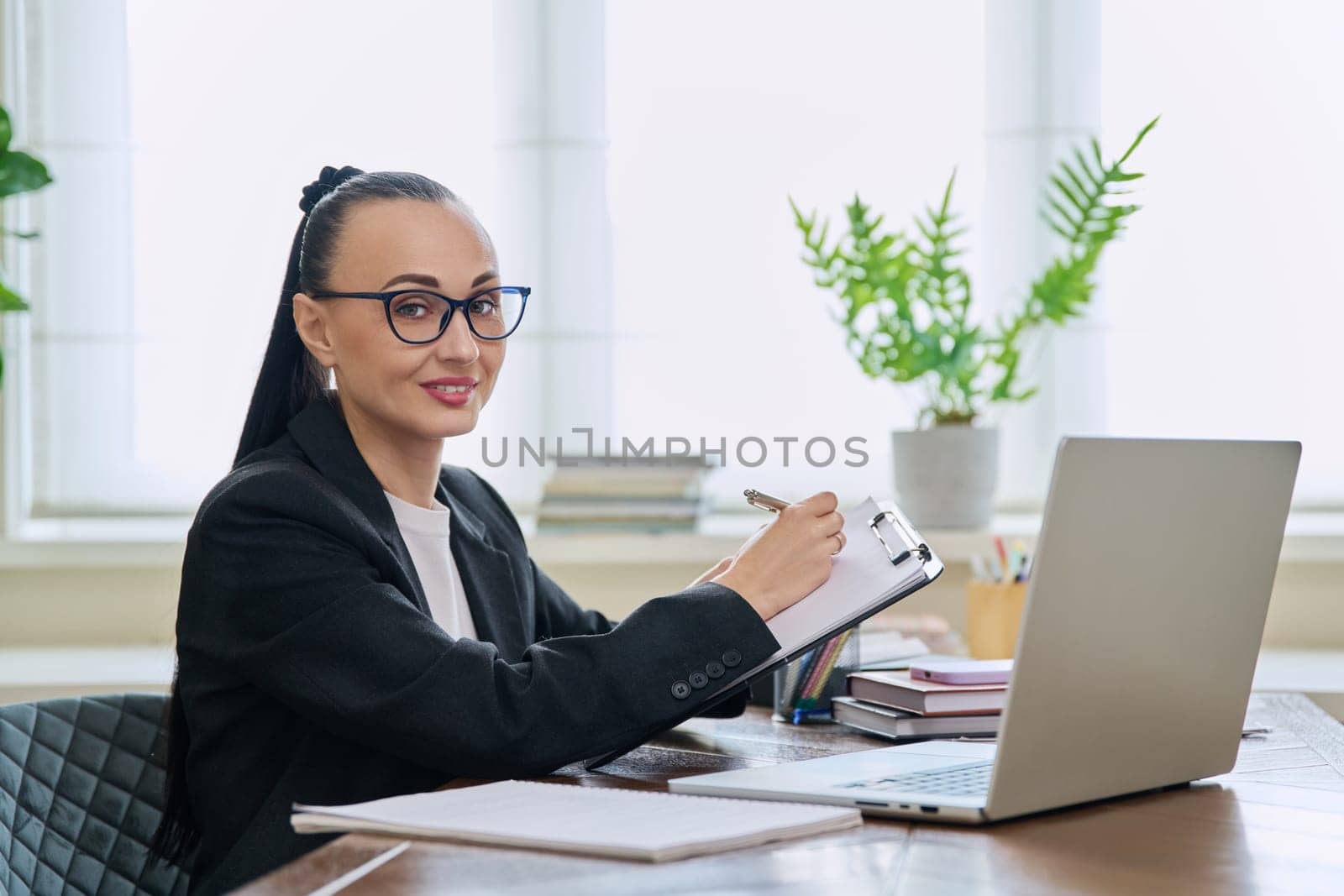 Portrait of businesswoman with clipboard sitting at workplace with computer laptop. Confident 30s female looking at camera. Remote business, technologies internet, teaching, blogging, work freelance