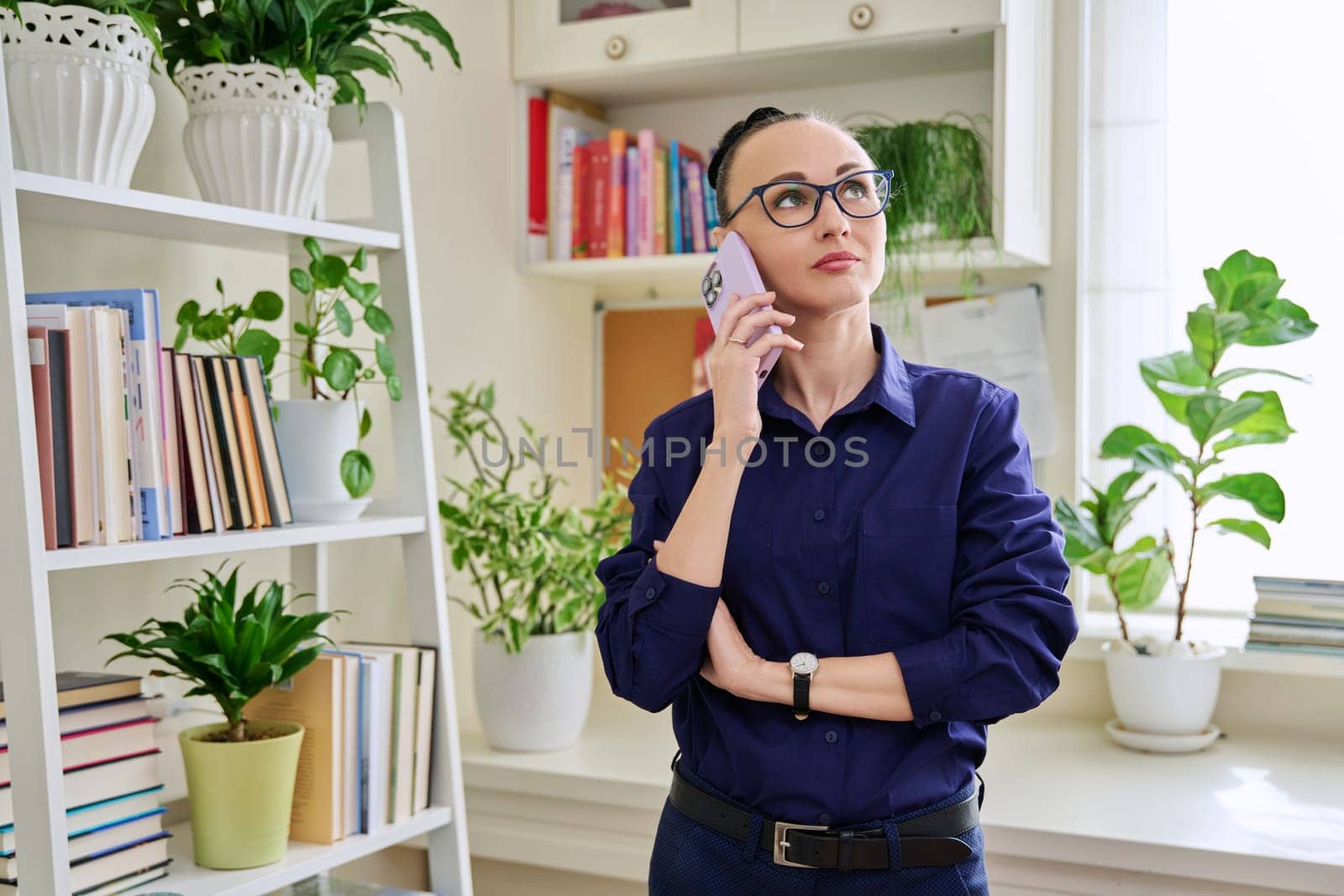 Beautiful emotional woman in her 30s talking on a cell phone, standing in her room at home by VH-studio