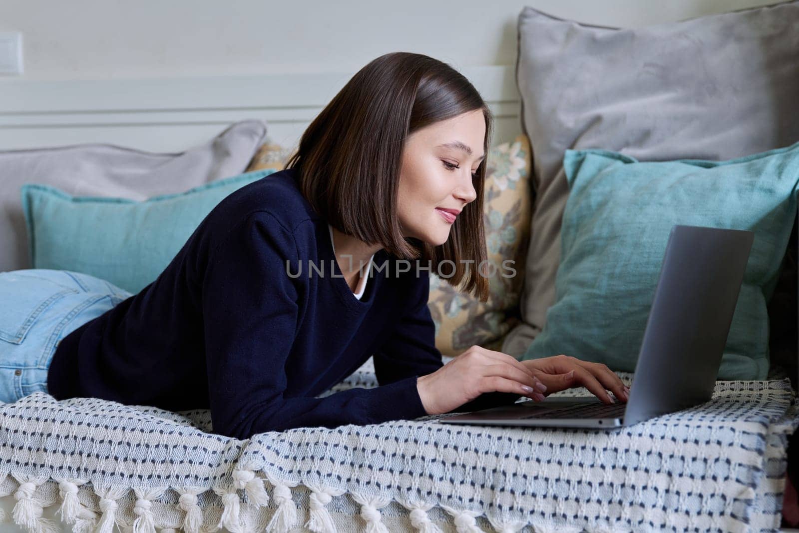 Young woman using laptop computer, typing on keyboard, lying on sofa at home. Internet online technologies for work communication study leisure