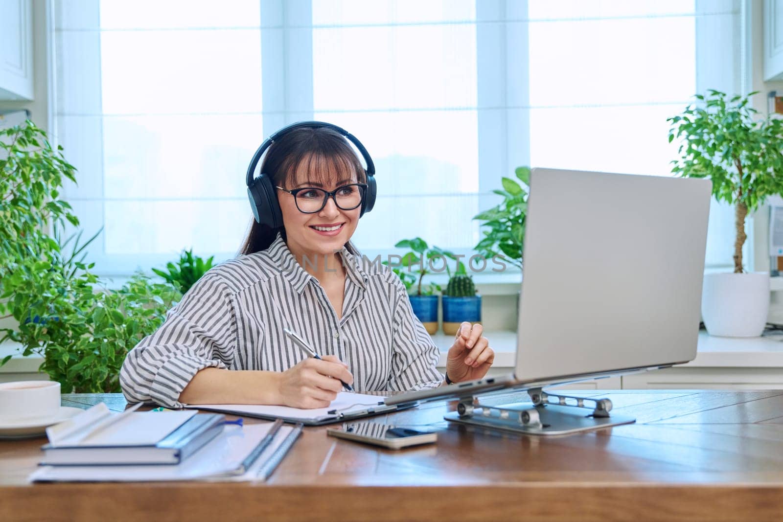 Mature woman in headphones having video conference using laptop computer in home office. Remote virtual meeting, online training, consulting, testing. Work, education, technology, people concept