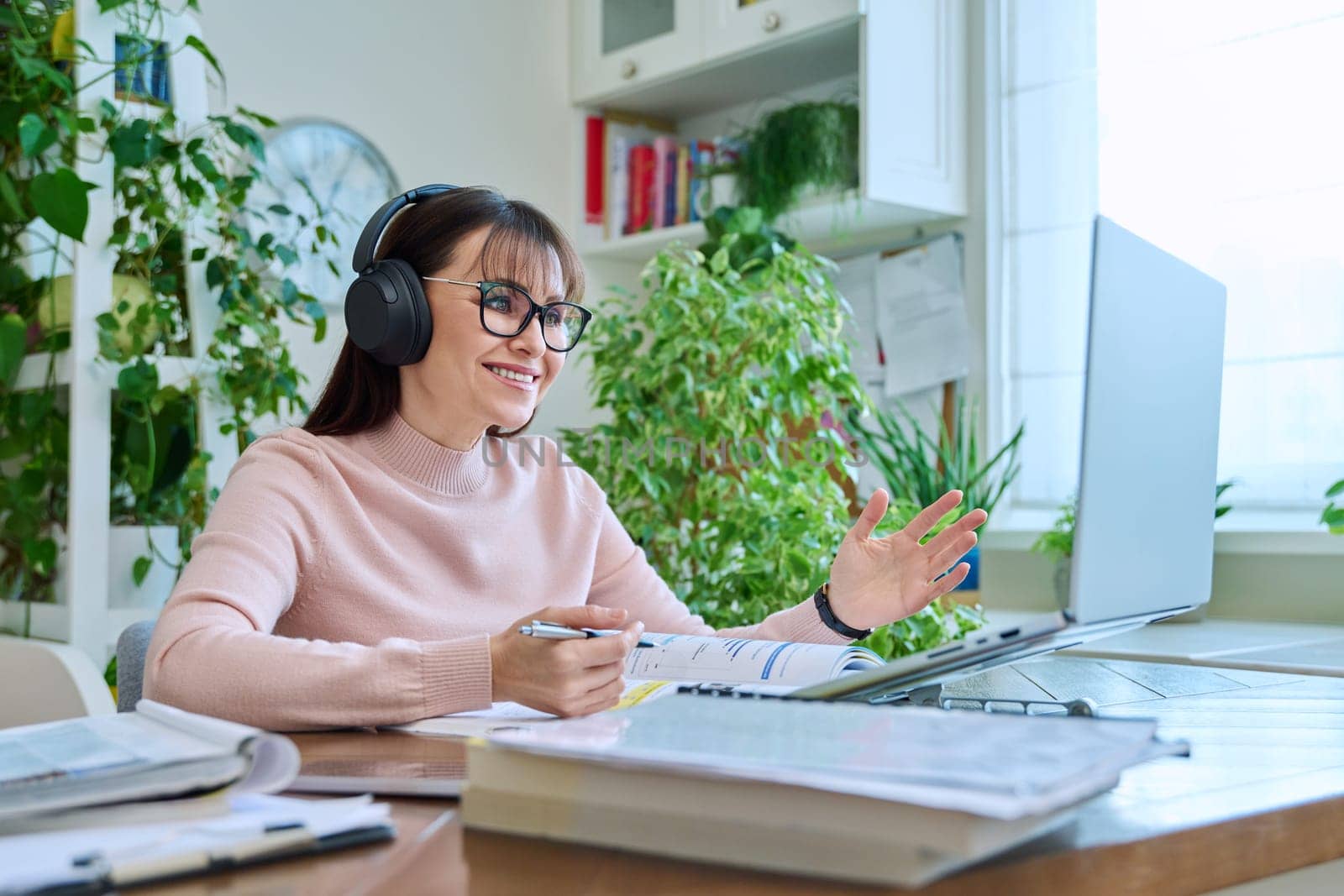 Middle aged woman teacher in headphones with textbook working online using laptop computer in home office. Video conference remote meeting consultation e lesson training education technology concept
