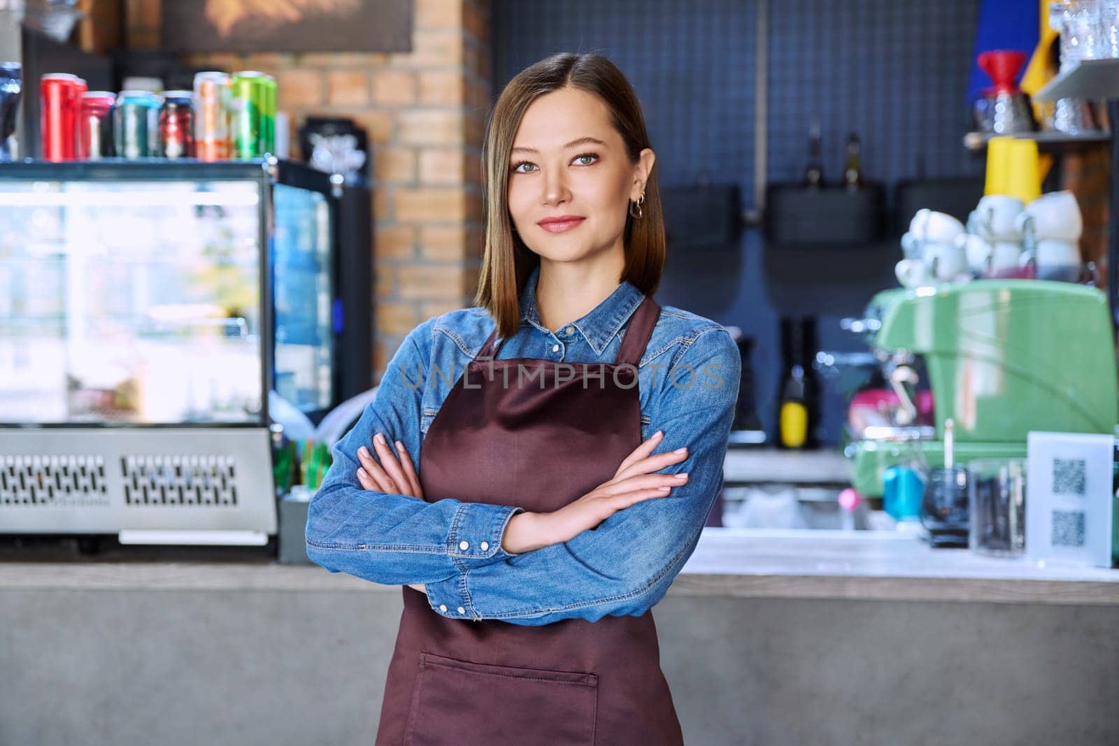 Young woman service worker in apron looking at camera in restaurant, coffee shop by VH-studio