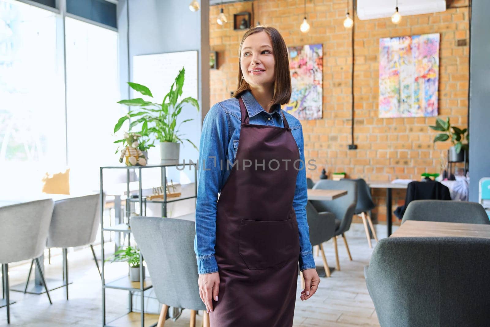 Young woman worker, owner in apron looking at camera in restaurant, coffee shop by VH-studio