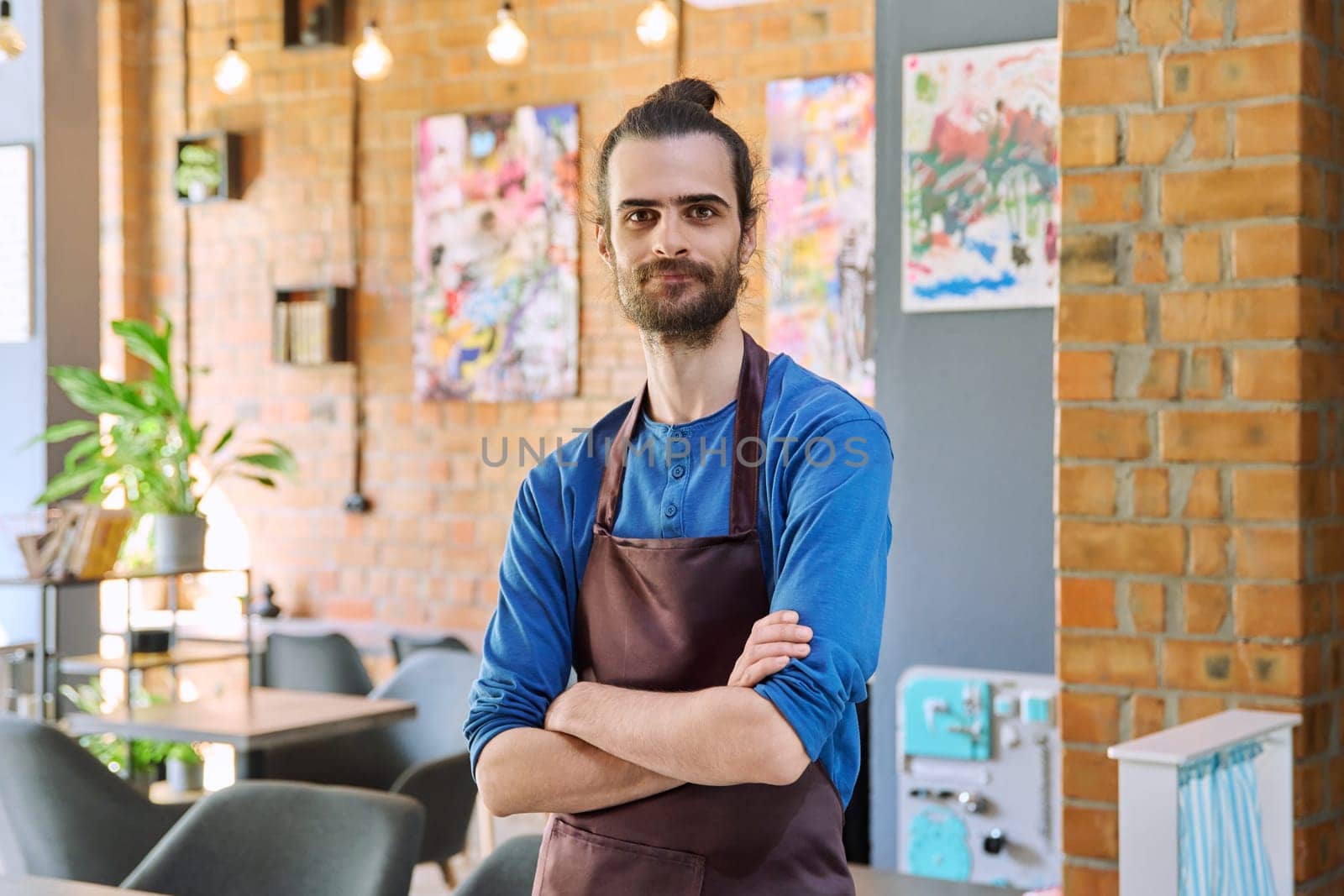Confident successful young man service worker owner in apron with crossed arms looking at camera in restaurant cafeteria coffee pastry shop interior. Small business staff occupation entrepreneur work