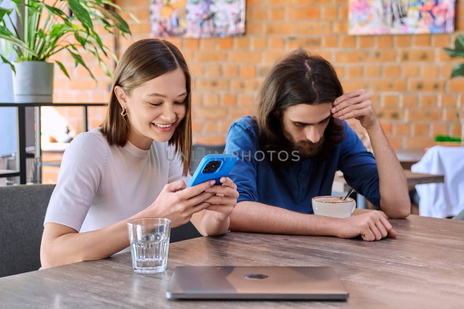 Young people, couple man and woman relaxing together in cafe, using smartphone by VH-studio