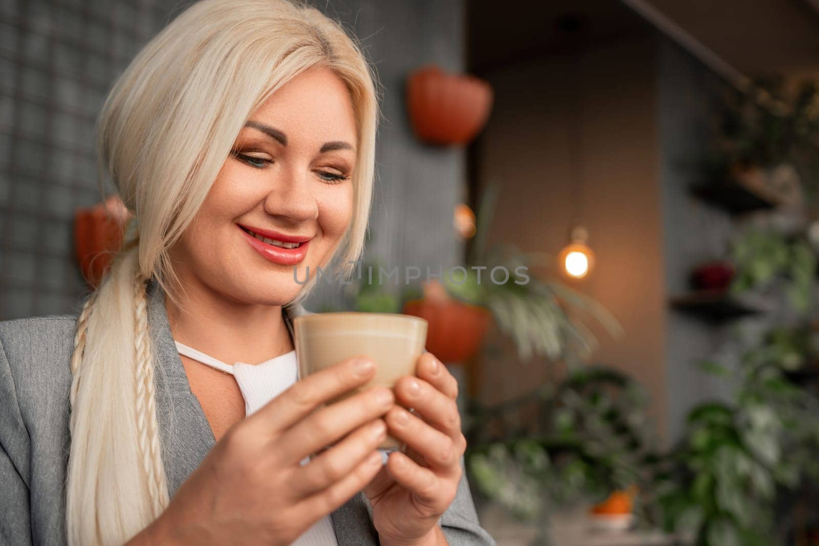 A blonde woman is holding a cup of coffee and smiling. The scene is set in a room with plants and a potted plant in the background