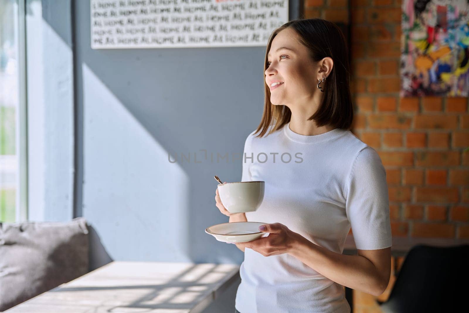 Young beautiful woman holding cup of coffee, looking out window, gray wall copy space by VH-studio