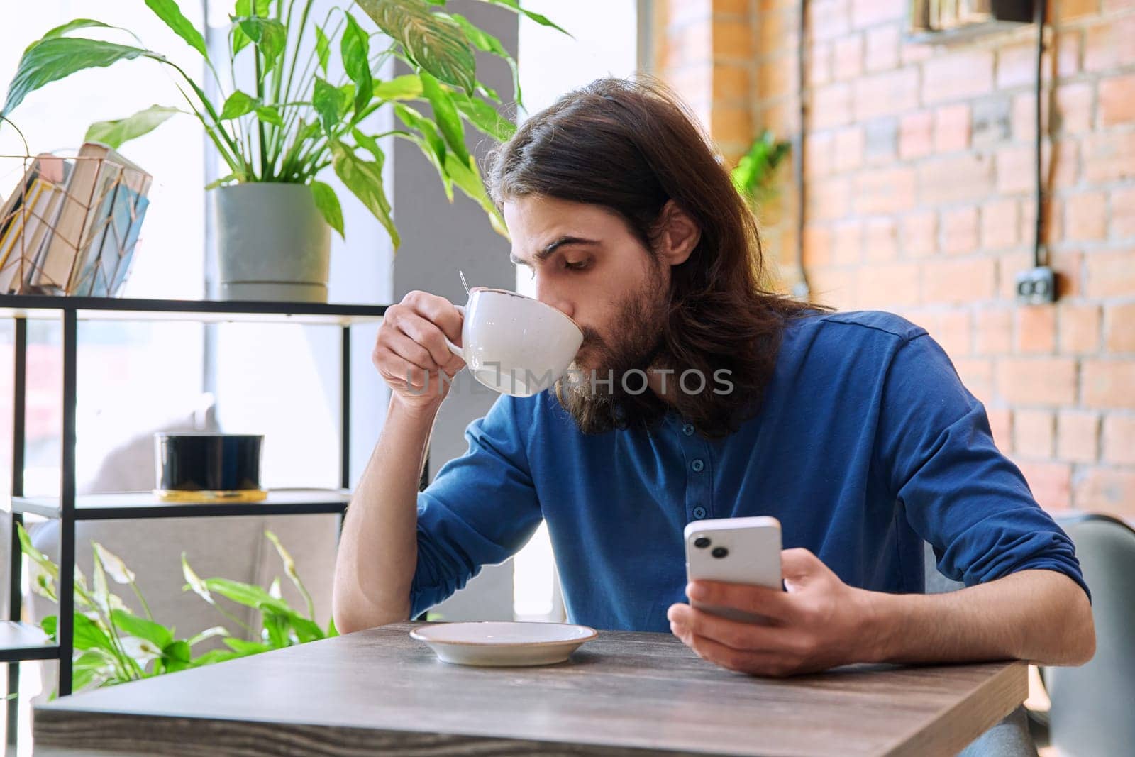 Young 30s stylish handsome bearded with long hair man using smartphone, drinking cup of coffee, sitting in cafe. Mobile technologies Internet apps applications for leisure work business communication