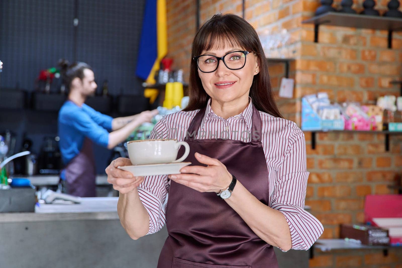 Woman in apron, food service coffee shop worker, small business owner with cup of fresh coffee, looking at camera near bar counter with male barista. Staff, occupation, entrepreneur, work concept