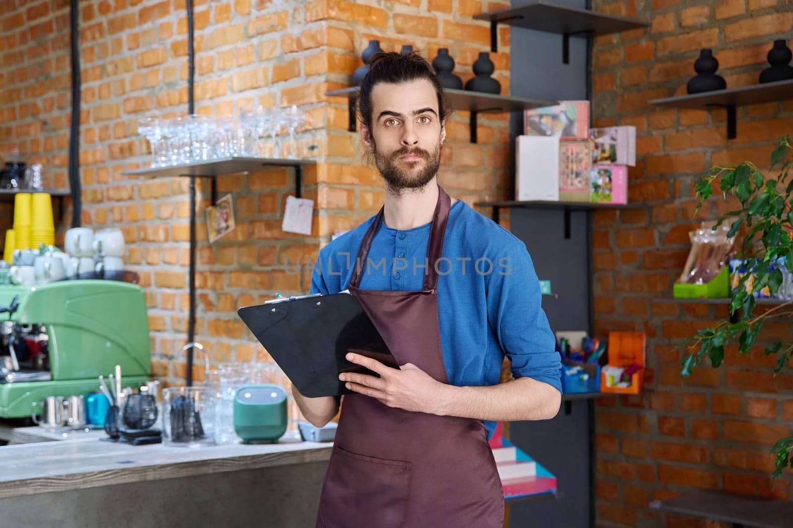 Young man in apron, food service worker, small business owner entrepreneur with work papers looking at camera near counter of coffee shop cafe cafeteria. Staff, occupation, successful business, work