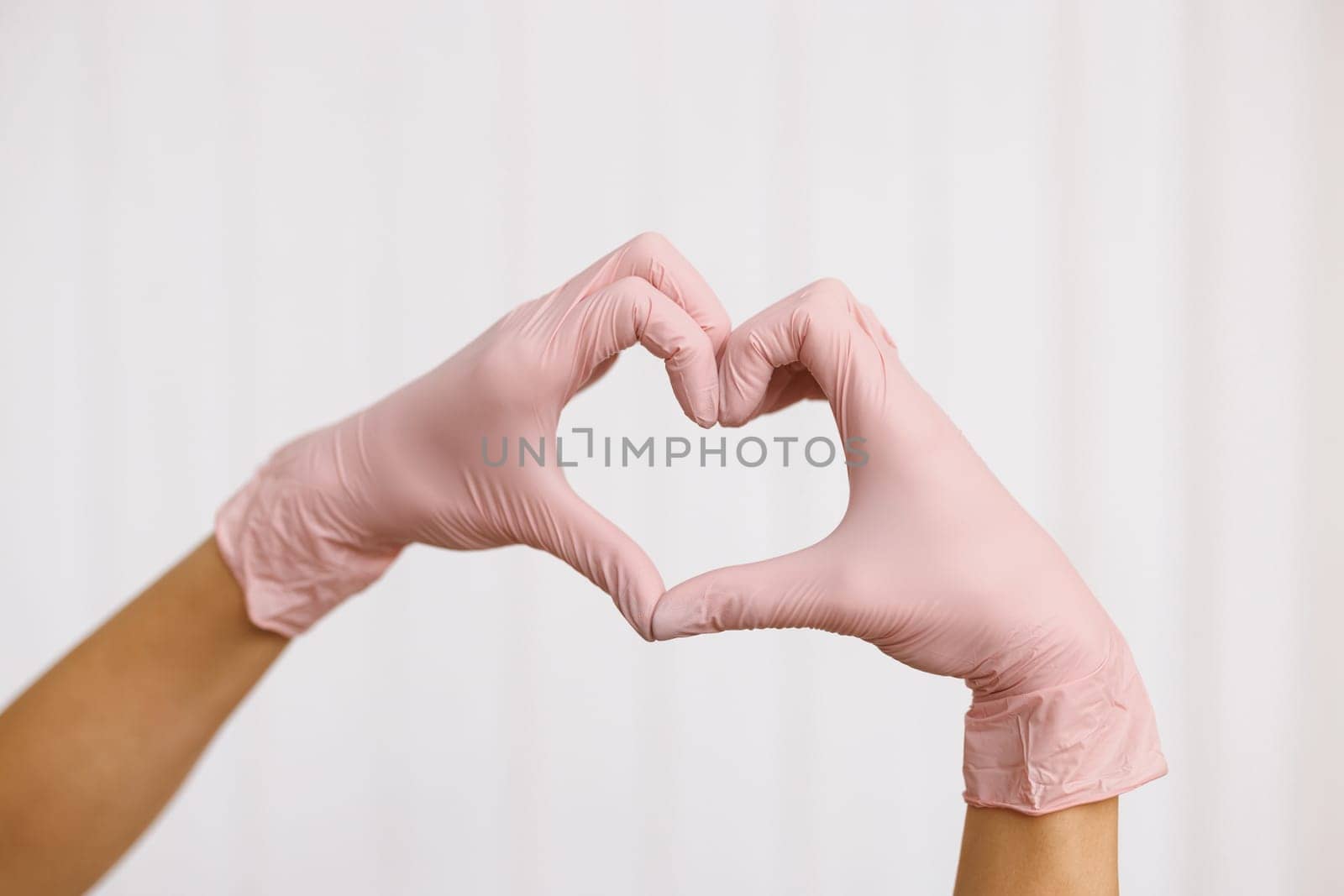 Person in pink latex gloves showing heart gesture against white background, closeup on hands. Hands with gloves making heart shape symbolize love or support to medical team by uflypro