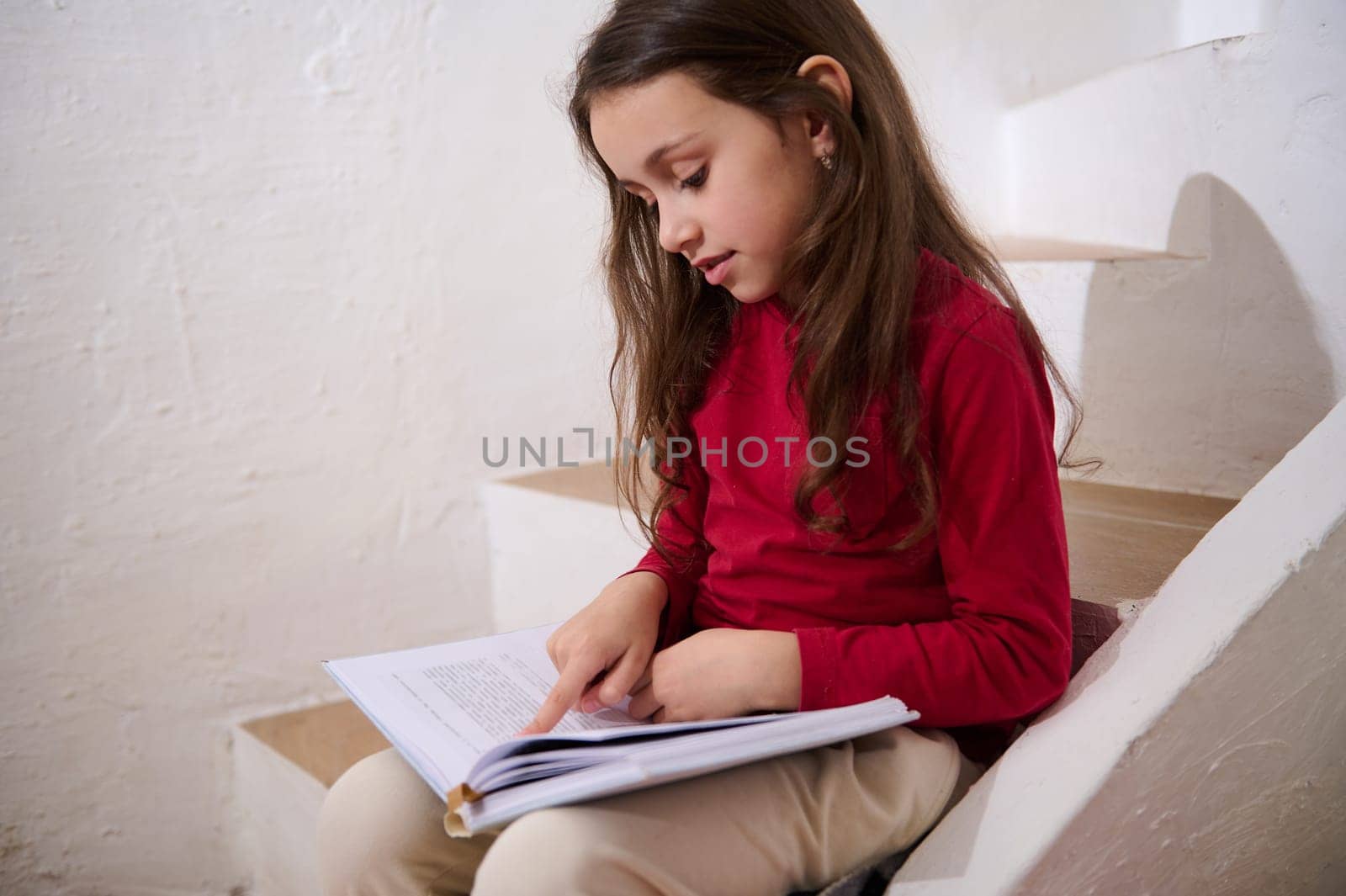 Caucasian cute child, elementary age smart little kid girl leafing through the pages of a hardcover book, reading a book, sitting on the steps at home, over white wall background of a rural house. by artgf