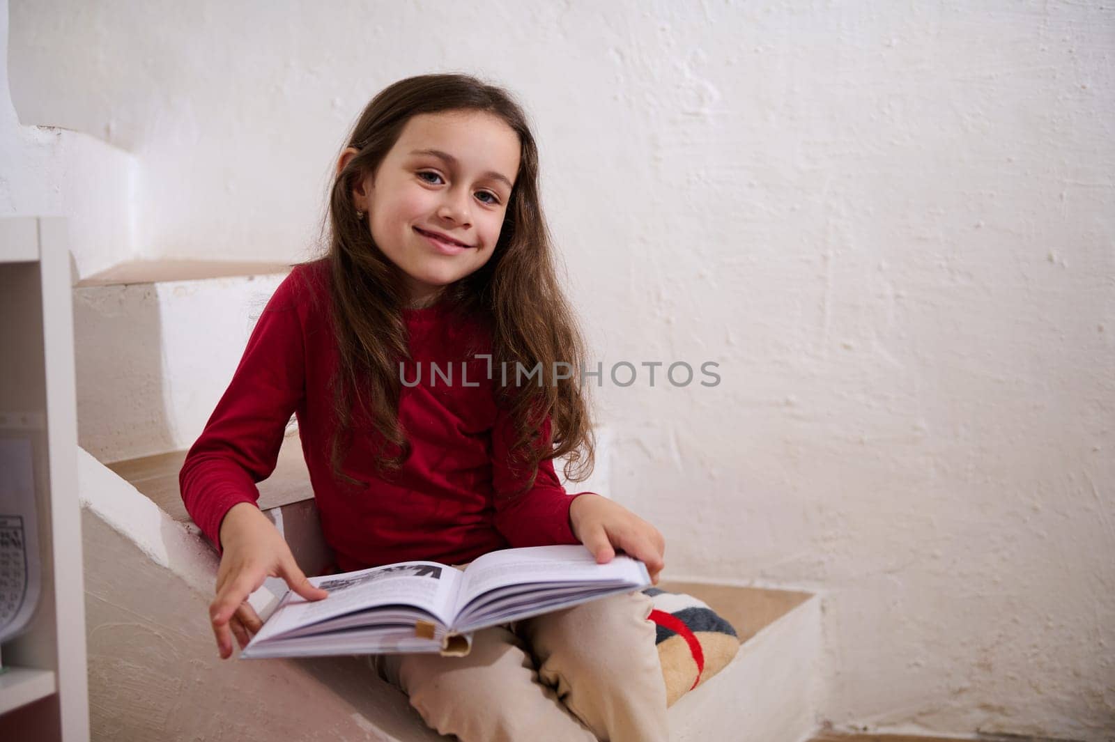 Smart primary school student girl smiling at camera, sitting on steps at home with a book in hands. Little girl reading a book. World Book Day concept. Back to school on new semester of academic year by artgf