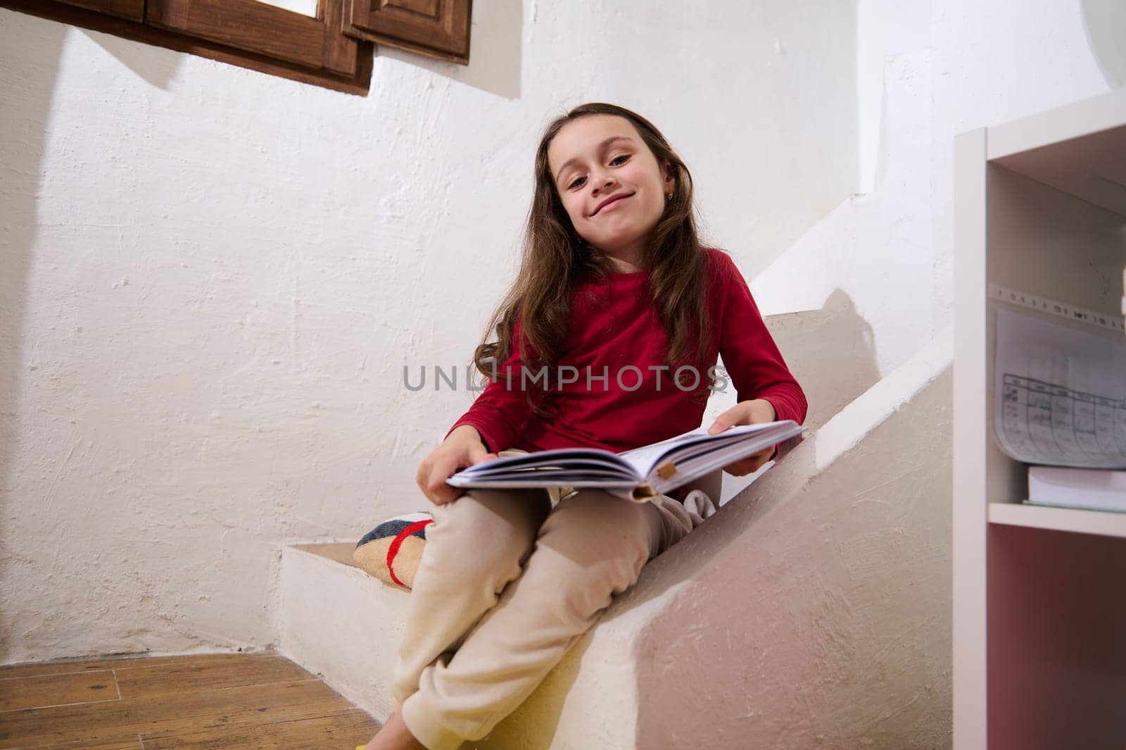 Adorable smart primary school student girl smiling looking at camera, sitting on steps at home with a book in hands. Intelligent little girl reading a book. World Book Day concept by artgf