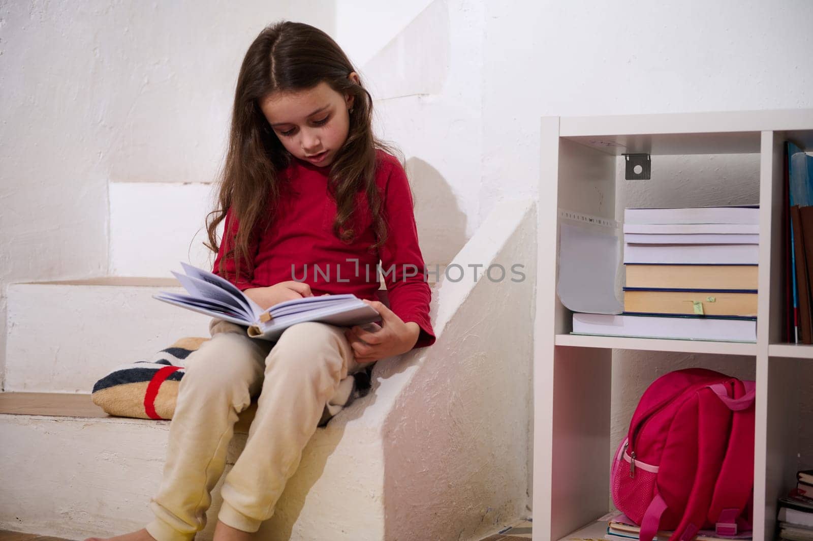 Cute school age child girl reading a book in cozy home atmosphere, sitting on steps of her room. The concept of back to school. Smart kids, Erudition and learning. World Book's Day by artgf
