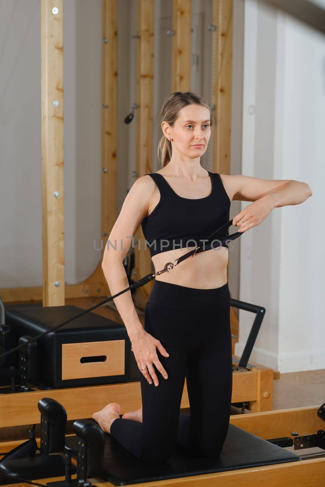 A young girl is doing Pilates on a reformer in the room. Beautiful slim woman in black clothes performs exercises to strengthen the spine and back muscles by Lobachad