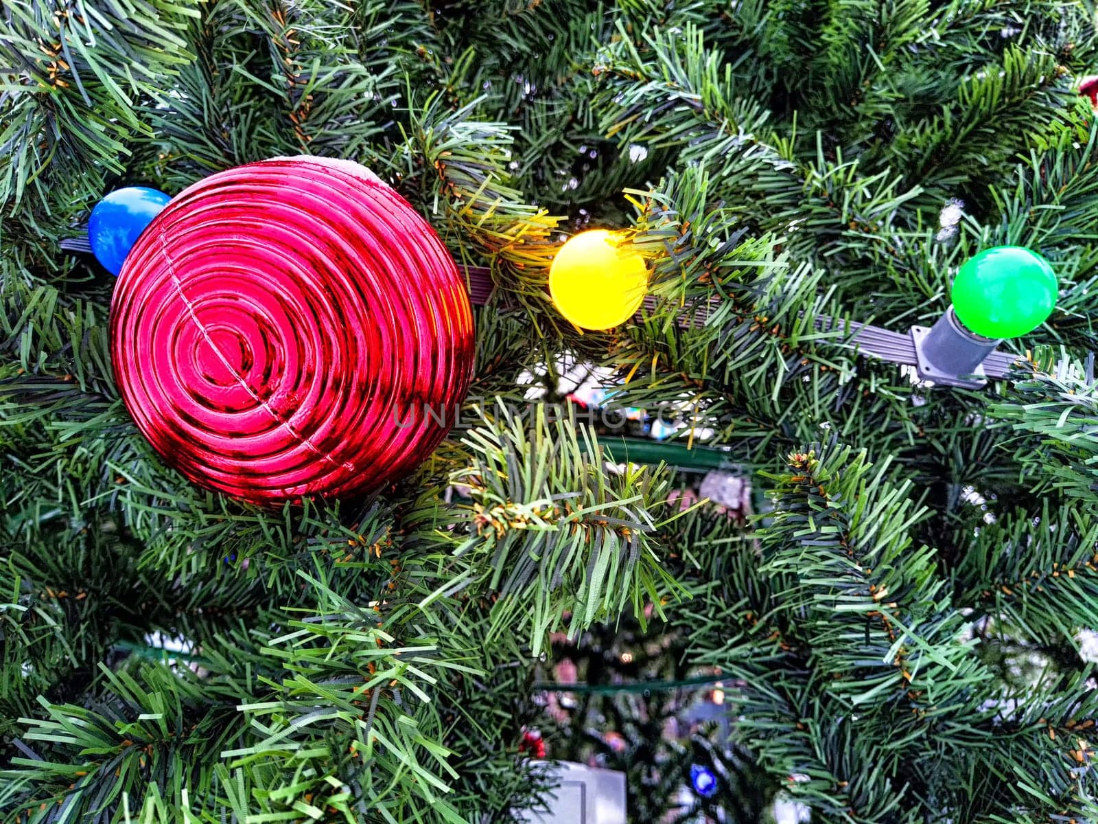 Festive Red Ornament Adorning a Lush Christmas Tree. A vibrant red ball nestled among green branches with multicolored lights by keleny