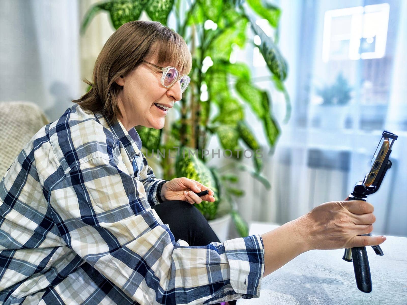 Smiling woman in glasses using a smartphone on a tripod for a video chat. A middle-aged woman posing and taking a selfie