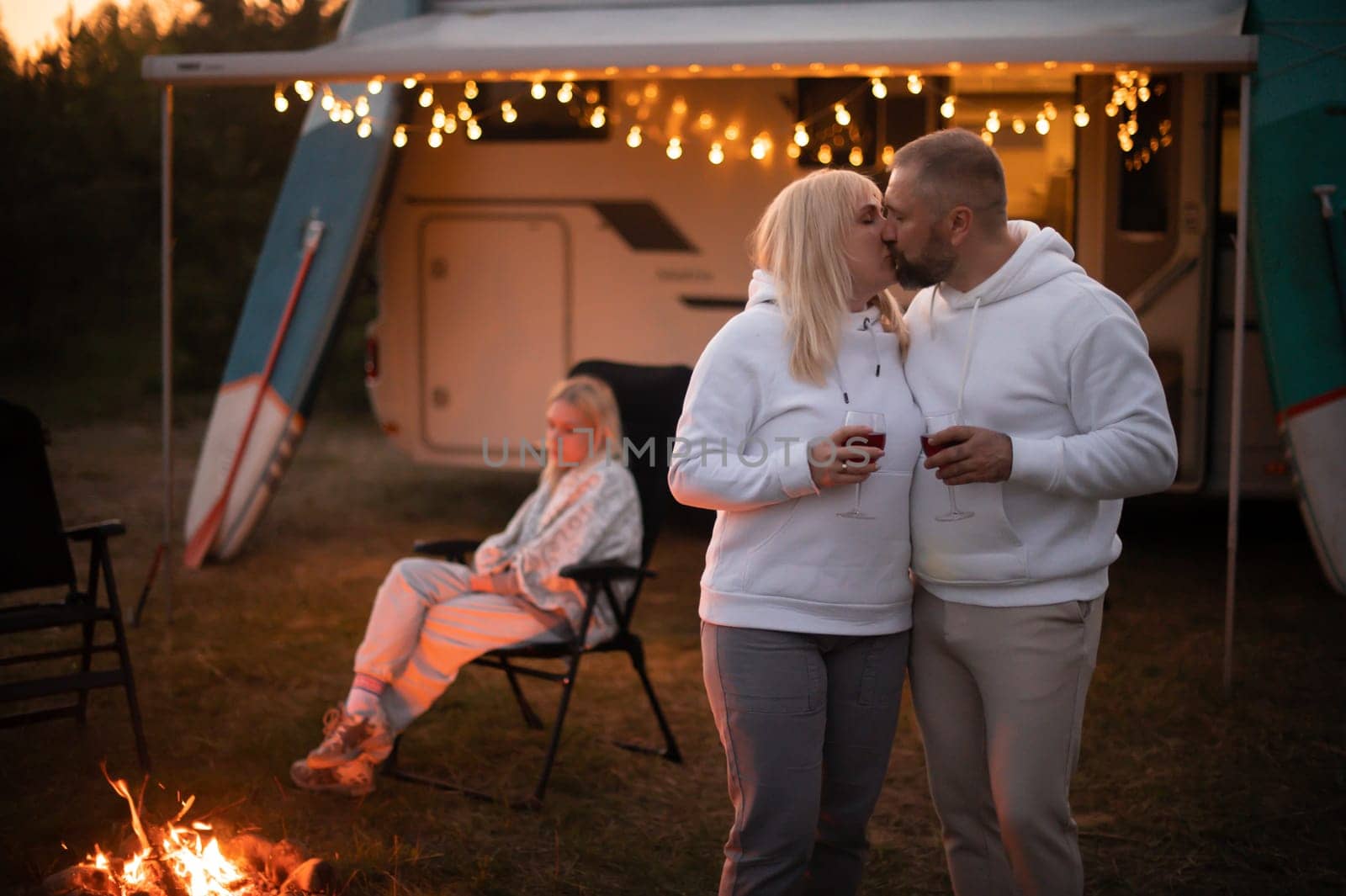 A married couple with glasses of wine stands against the background of a motorhome and rests together by the campfire. Evening family vacation.