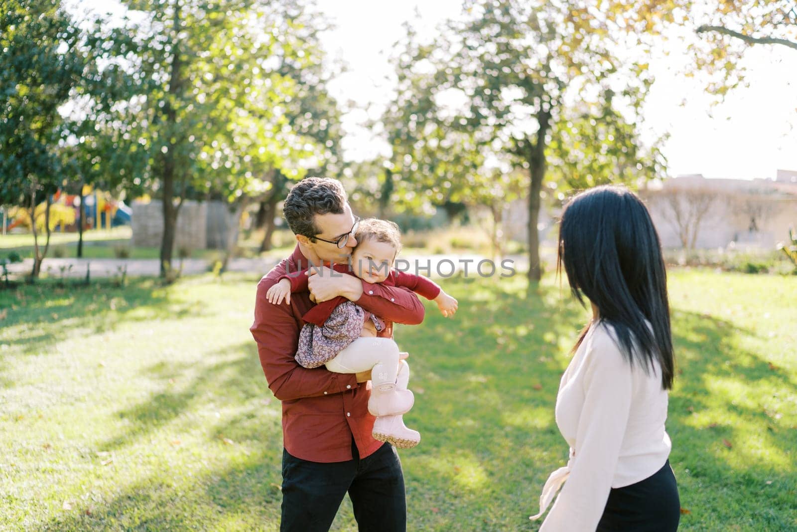 Dad kisses a little girl in his arms next to a standing mom in the park. High quality photo