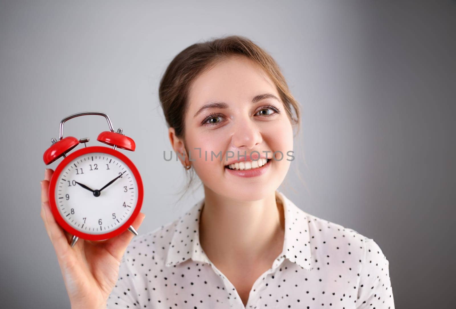 Adult smiling caucasian woman hold red alarm clock on gray background.