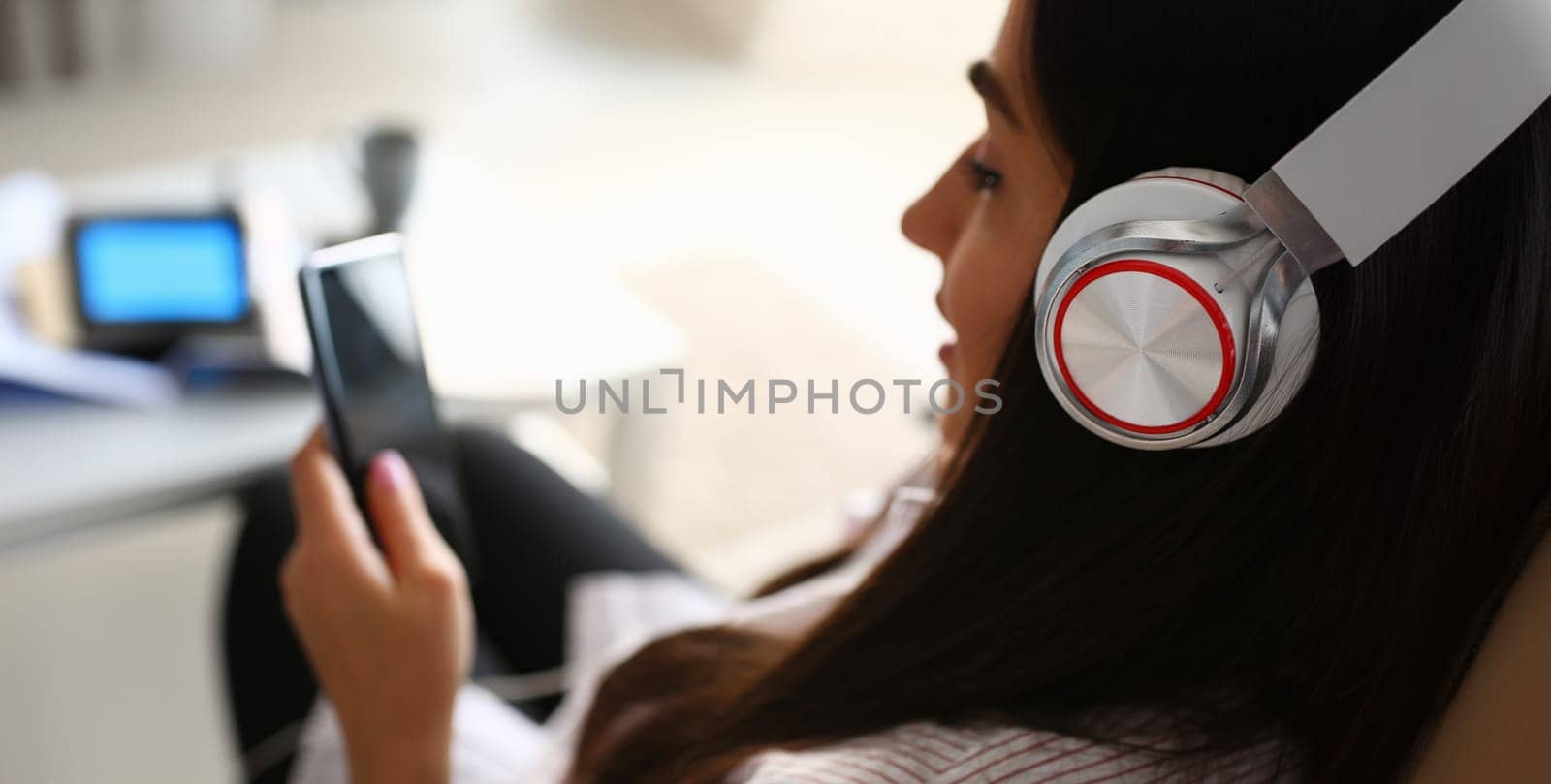 Young beautiful woman headphones holds smartphone in hand using at home costs about sofa. Verifies internal information its financial advertising freedom makes money level transfer contractor concept