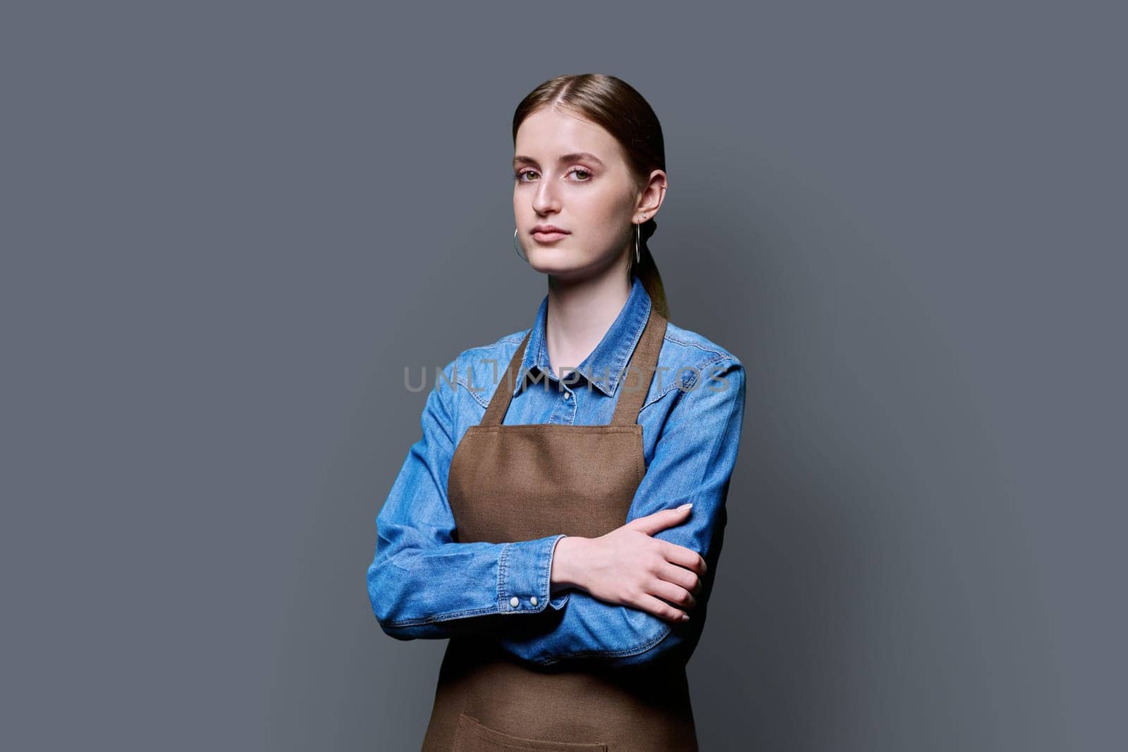 Portrait of young serious confident woman in apron on grey background by VH-studio