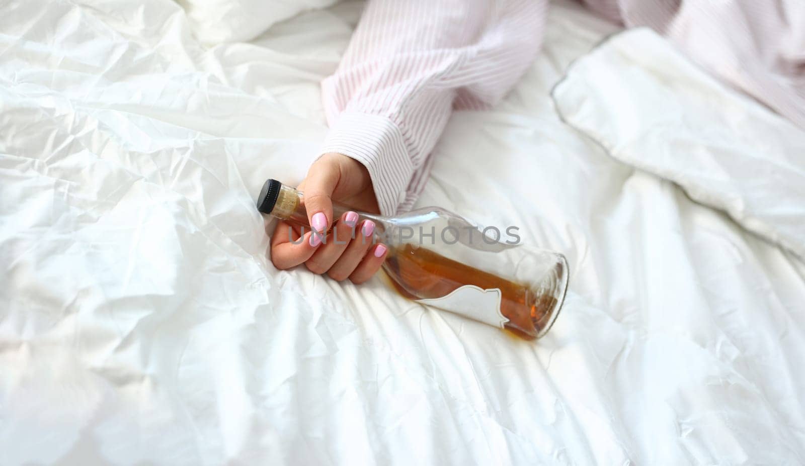 Young woman lying in bed deadly drunken holding near-empty bottle of booze. Female intoxicated with alcohol after tough night party. Alcoholism habitual drunkenness pernicious habit concept