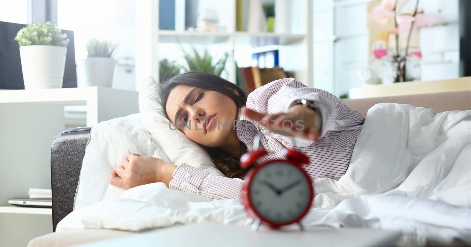 Sleepy young woman portrait with one opened eye trying kill alarm clock. Early wake up not getting enough sleep going work concept. Female stretching hand to ringing alarm willing turn it off