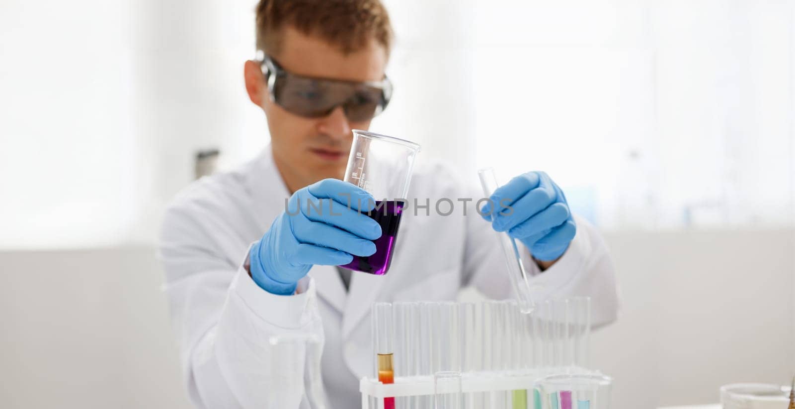 Male chemist holds test tube of glass in his hand overflows a liquid solution of potassium permanganate conducts an analysis reaction takes various versions of reagents using chemical manufacturing.