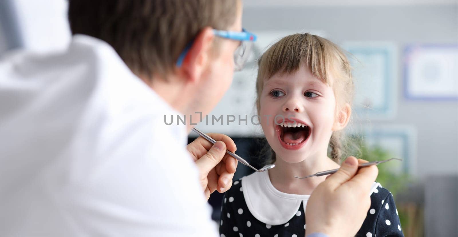 Male dentist look at open mouth litle happy girl child againlt hospital office background portrait. Tooth treatment concept