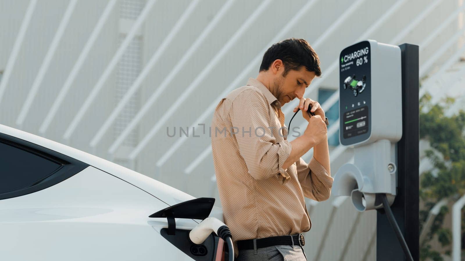 Young man put EV charger to recharge electric car's battery from charging station in city commercial parking lot. Rechargeable EV car for sustainable environmental friendly urban travel. Expedient