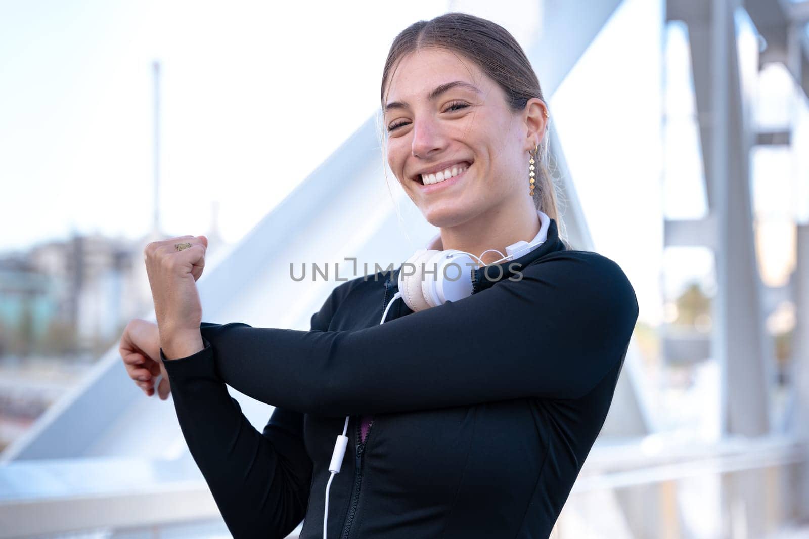 Young happy sportswoman in sports headphones doing stretching smiling looking at camera outdoors.