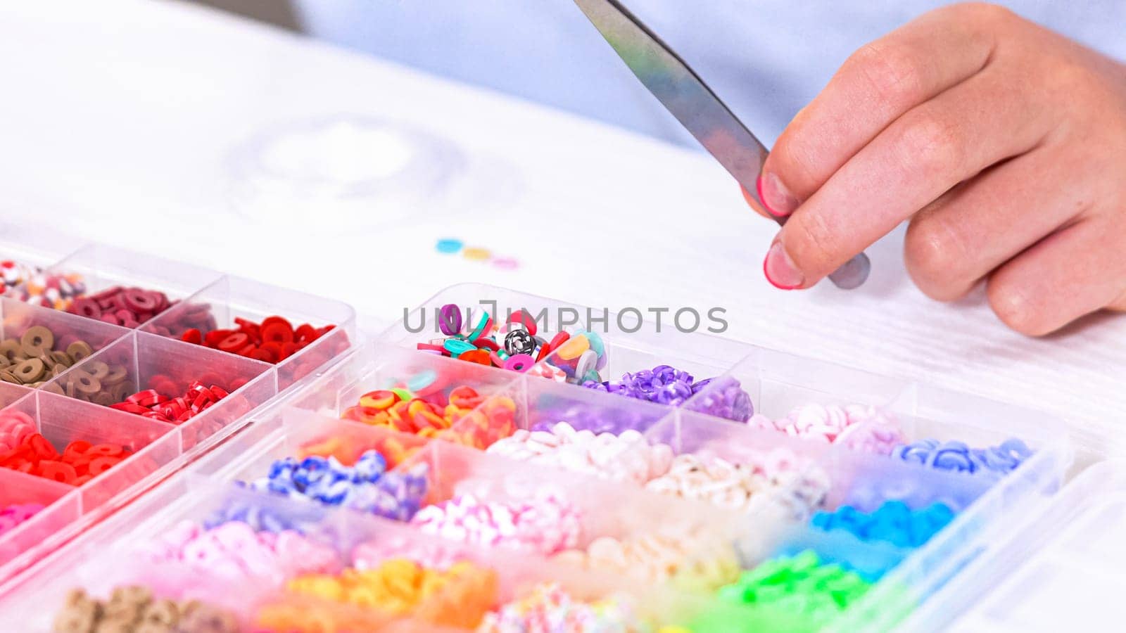 Delicate fingers of a young girl navigate through a treasure trove of bright, multicolored beads, each compartment revealing a new hue to choose from. She's immersed in the joyful task of stringing together a handmade bracelet, with golden beads and pearls lying nearby to add a touch of sparkle to her creation.