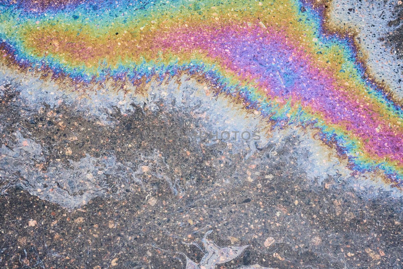 Close-up of an iridescent oil or gasoline spill on a wet asphalt, viewed from above. Bold multicolored spots on the asphalt.