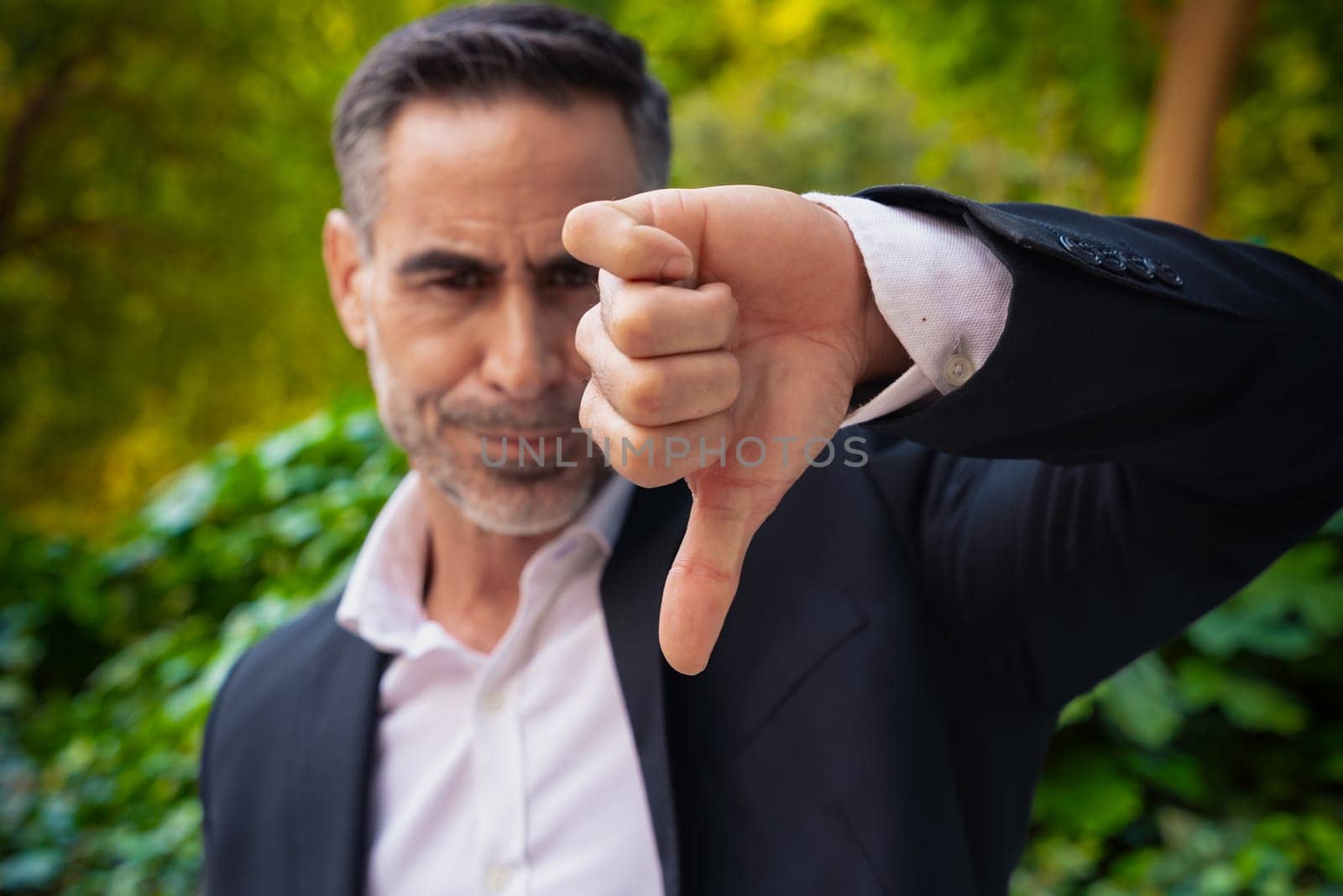 Serious businessman with thumbs down looking at camera outdoors. by mariaphoto3