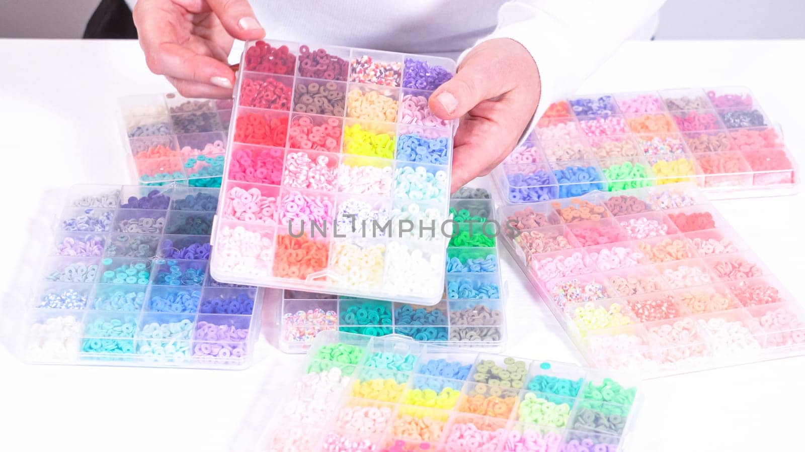 Woman hands gracefully poised over a collection of beads, sorted by color in transparent organizers. The array of beads spans a vibrant spectrum, from deep purples to bright oranges, meticulously arranged for easy selection as she embarks on creating a custom piece of jewelry.