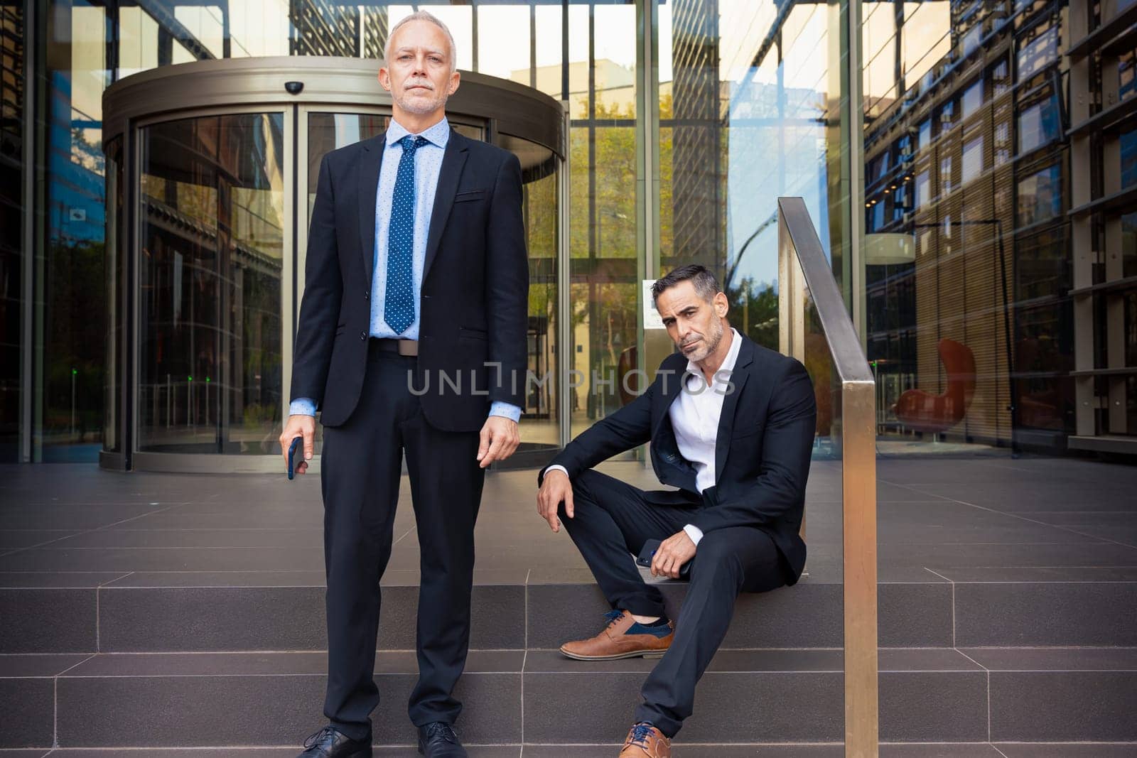 Portrait Of Two Serious Trusted Business Colleagues Dressed In Formal Suit One Standing And The Other Sitting, Together Outside The Workplace During Work Meeting