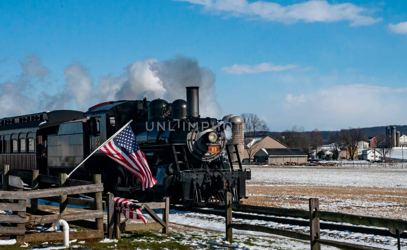 Ronks, Pennsylvania, USA, February 17, 2024 - A steam train is pulling a train car with an American flag on it. The train is traveling through a snowy field