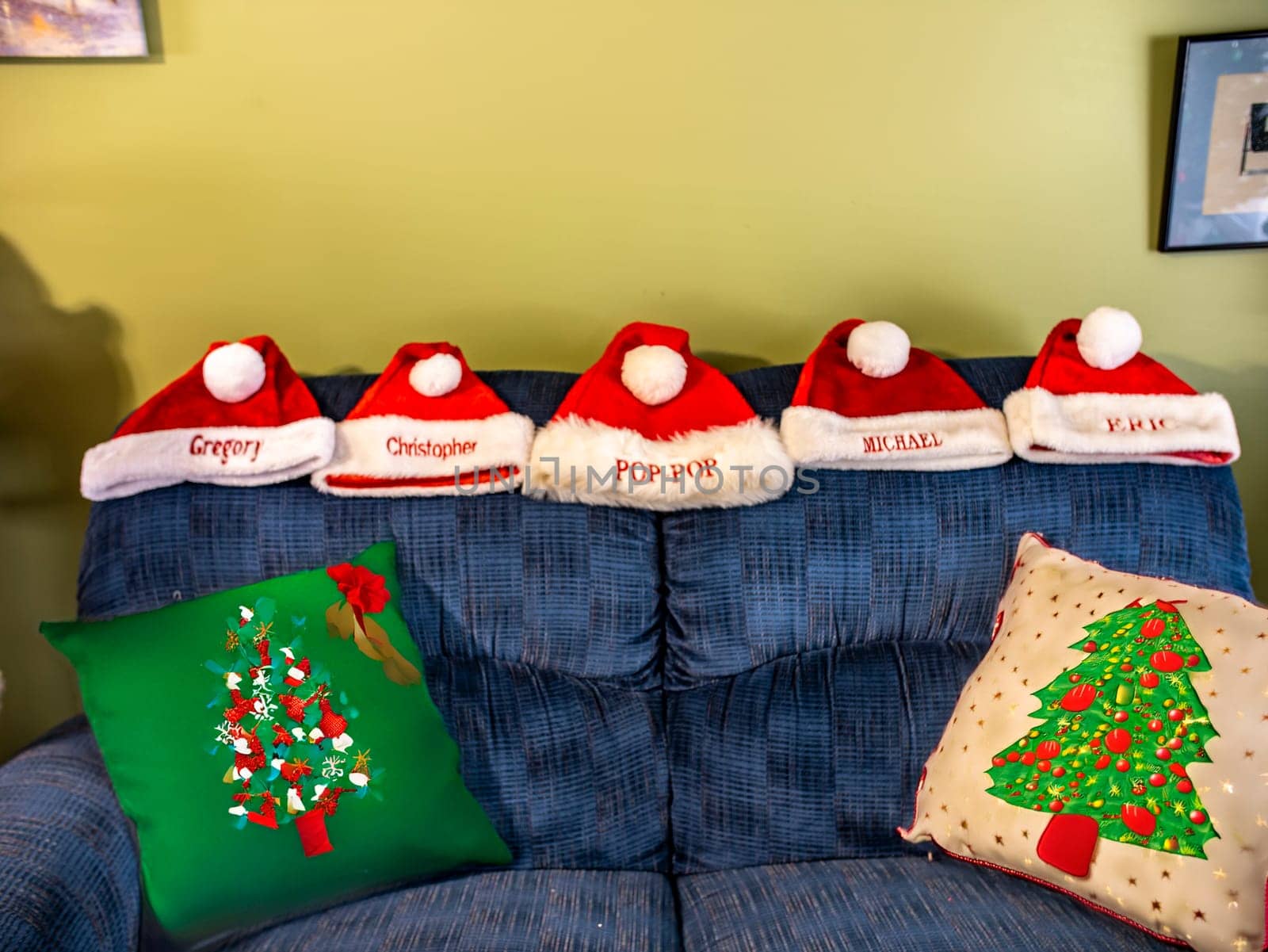 A group of five red santa hats are sitting on a blue couch. The hats are personalized with names and are arranged in a row. The couch is decorated with pillows